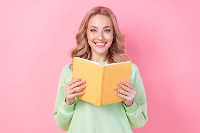 ピンクの背景に立つ黄色い本を持った女性